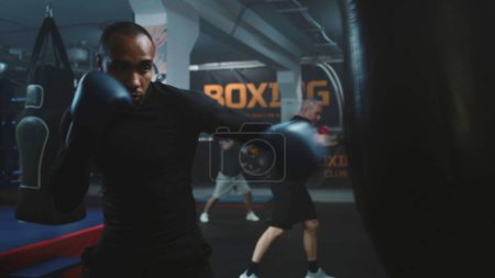 Photo for African American fighter listens coach command and starts hitting punching bag in dark boxing gym. Athletic man in boxing gloves exercises before tournament. Physical activity and intensive workout. - Royalty Free Image