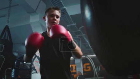 Photo for Confident young fighter hits punching bags while training in dark boxing gym. Athletic boy in boxing gloves exercises with trainer before fighting tournament. Physical activity and intensive workout. - Royalty Free Image