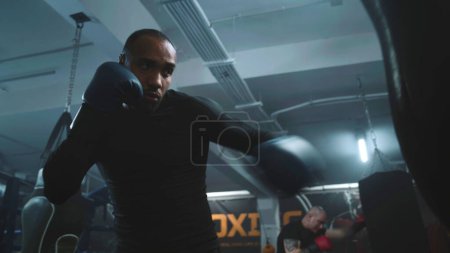 Photo for African American fighter listens coach command and starts hitting punching bag in dark boxing gym. Athletic man in boxing gloves exercises before tournament. Physical activity and intensive workout. - Royalty Free Image