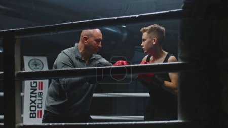 Photo for Professional coach teaches young boxer how to dodge punches on boxing ring. Athletic boy hits punching mitts while training before fighting competition. Physical activity and intensive workout concept - Royalty Free Image