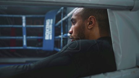 Photo for Tired and exhausted African American boxer sits in boxing ring corner. Fighter with beads of sweat on face rests after training. Athlete prepares to tournament or competition in boxing gym. Close up. - Royalty Free Image
