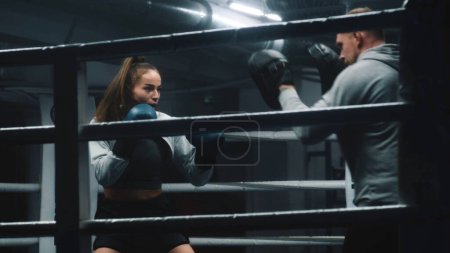 Photo for Fighter in boxing gloves practices fighting technique with male trainer and exercises before competition in dark gym. Athletic woman hits punching mitts on boxing ring. Physical activity and workout. - Royalty Free Image