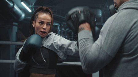 Photo for Close up of female fighter in boxing gloves practicing fighting technique and exercising before match in gym. Athletic boxer hits punching mitts on boxing ring. Physical activity and workout concept. - Royalty Free Image