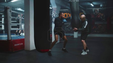 Téléchargez les photos : Boxer afro-américain en gants de boxe exercices avec entraîneur dans le gymnase sombre avant la compétition de combat. Homme sportif frappe sac de boxe tout en pratiquant dans le gymnase de boxe. Entraînement de combat en salle de gym. - en image libre de droit