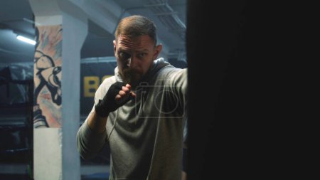 Photo for Close up of male fighter training and preparing for fighting match in dark boxing gym. Athletic man in boxing bandages hits and practices fighting technique. Physical activity and intensive workout. - Royalty Free Image