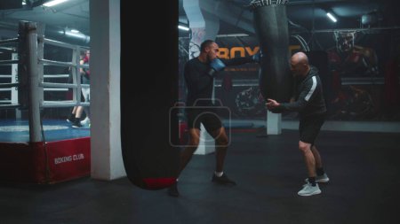 Photo for African American boxer in boxing gloves exercises with trainer in dark gym before fighting competition. Athletic man hits punching bag while practicing in boxing gym. Fighter training in box gym. - Royalty Free Image