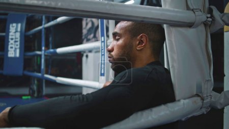 Téléchargez les photos : Boxer afro-américain fatigué et épuisé est assis dans un coin de boxe. Combattant avec des perles de sueur sur le visage repose après l'entraînement. Athlète se prépare au tournoi ou à la compétition dans la salle de boxe. Gros plan. - en image libre de droit