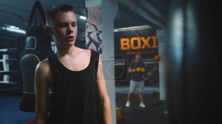 Photo for Confident young fighter hits punching bag while training in dark boxing gym. Athletic teenager in boxing gloves exercises before fighting tournament. Physical activity and intensive workout in gym. - Royalty Free Image