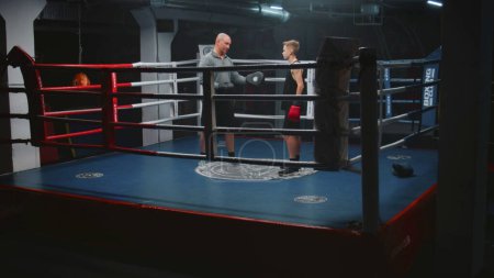 Photo for Boy in boxing gloves stands on ring and talks with trainer. Adult man consults young boxer and explains fighting techniques. Teen prepares to fighting match in dark gym. Physical activity and workout. - Royalty Free Image