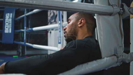 Photo for Tired and exhausted African American boxer sits in boxing ring corner. Fighter with beads of sweat on face rests after training. Athlete prepares to tournament or competition in boxing gym. Close up. - Royalty Free Image