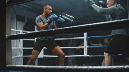 Téléchargez les photos : Boxer afro-américain dans les exercices de gants de boxe et se prépare pour la compétition de combat avec un entraîneur caucasien sur le ring. Homme combattant frappe mitaines de poinçonnage dans la salle de gym sombre. Activité physique et entraînement. - en image libre de droit