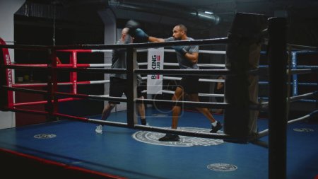 Téléchargez les photos : Des combattants afro-américains s'entraînent et se préparent pour un match avec un entraîneur caucasien sur un ring de boxe. Homme sportif en gants de boxe frappe mitaines de boxe dans la salle de gym sombre. Activité physique et entraînement. - en image libre de droit