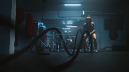 Photo for Female athlete exercises with battle ropes in dark boxing gym with LED lighting. Female boxer does cardio or endurance workout before championship fight. Physical activity and CrossFit training. - Royalty Free Image