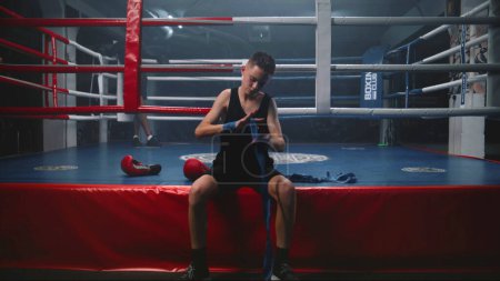 Photo for Young boxer sits near boxing ring and wraps his hands with bandage for martial arts. Teenage athlete prepares to fight tournament or competition, trains in boxing gym. Physical activity. Dolly shot. - Royalty Free Image