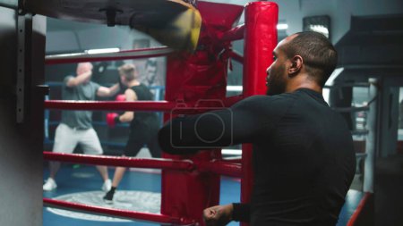 Téléchargez les photos : Un combattant afro-américain concentré frappe un sac de boxe pendant l'entraînement dans un gymnase de boxe sombre. Exercices d'homme athlétique avant la compétition ou le tournoi de combat. Boxer se prépare à se battre et s'entraîne. Vue arrière. - en image libre de droit