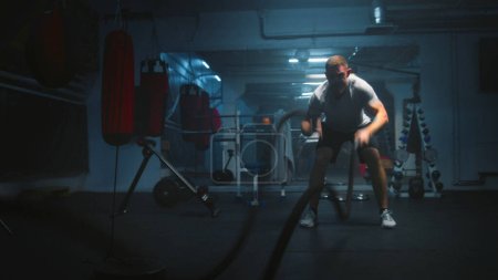 Photo for Male athlete exercises with battle ropes in dark boxing gym with LED lighting. Professional boxer does cardio or endurance workout before championship fight. Physical activity and CrossFit training. - Royalty Free Image