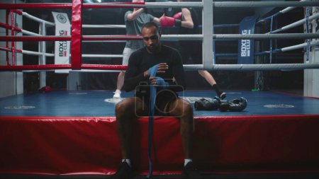 Photo for African American boxer sits near boxing ring and wraps hands with bandage for martial arts. Trainer practices punches with teenager at background. Physical activity and intensive workout. Dolly shot. - Royalty Free Image