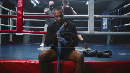 Photo for African American boxer sits near boxing ring and wraps hands with bandage for martial arts. Trainer practices punches with teenager at background. Physical activity and intensive workout. Dolly shot. - Royalty Free Image