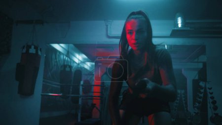 Photo for Athletic woman trains with battle ropes in dim boxing gym with LED lighting. Professional female boxer does cardio or endurance exercises before fighting competition. Intensive CrossFit workout. - Royalty Free Image