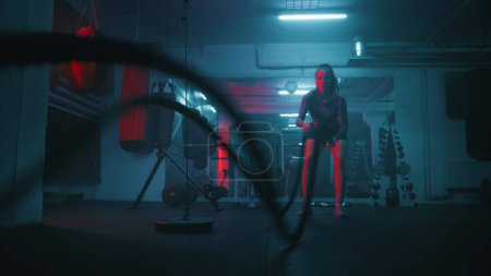 Photo for Female athlete exercises with battle ropes in dark boxing gym with LED lighting. Female boxer does cardio or endurance workout before championship fight. Physical activity and CrossFit training. - Royalty Free Image