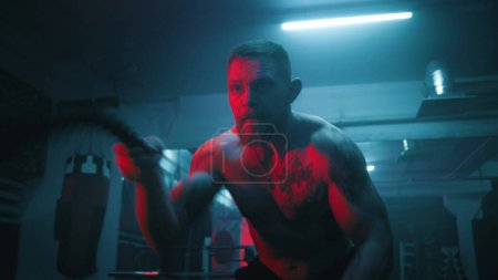 Photo for Male athlete exercises with battle ropes in dark boxing gym with LED lighting. Professional boxer does cardio or endurance workout before championship fight. Physical activity and CrossFit training. - Royalty Free Image