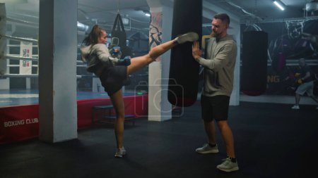 Téléchargez les photos : Femme kickboxer dans les bandages de boxe frappe sac de boxe lors de l'entraînement dans le gymnase de boxe sombre. Athlétique femme pratique et se prépare pour le match avec l'entraîneur masculin. Activité physique et entraînement intensif. - en image libre de droit