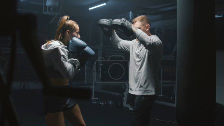 Photo for Athletic fighter in boxing gloves trains and exercises for fighting championship. Woman hits punching mitts and practices in fighting techniques with adult trainer. Physical activity and workout. - Royalty Free Image