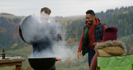 Photo for Caucasian man takes meat out of the barbecue grill for friends and kids. Multiethnic group of tourists or big family resting outdoors during vacation trip in the mountains. Tourism and active leisure. - Royalty Free Image