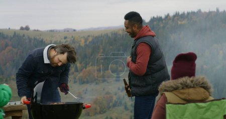 Photo for Caucasian man takes meat out of the barbecue grill for friends and kids. Multiethnic group of tourists or big family resting outdoors during vacation trip in the mountains. Tourism and active leisure. - Royalty Free Image