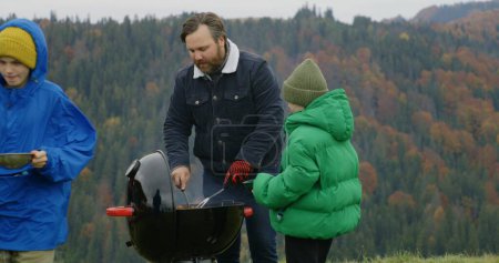 Photo for Adult man takes meat out of the barbecue grill for children. Multiethnic group of travelers or big family resting outdoors during vacation trip. Mountains landscapes and forest in the background. - Royalty Free Image