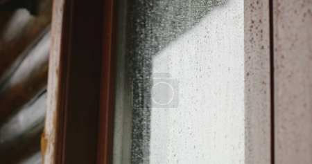 Foto de Primer plano de gotas de lluvia corriendo hacia abajo en la superficie de vidrio nebulizado de la ventana fuera de la casa en el campo o las montañas. Clima ventoso, lluvioso y húmedo en otoño al aire libre. Belleza de la naturaleza. - Imagen libre de derechos