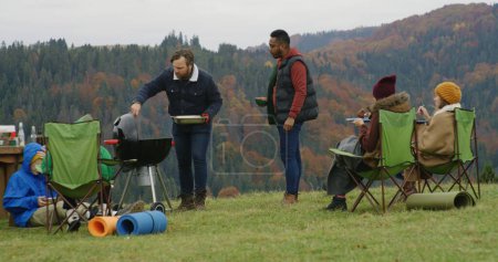 Photo for Caucasian man cooks in barbecue grill on the hill for friends and kids. Big multiethnic family of tourists eat and rest outdoors during vacation trip in the mountains. Tourism and active leisure. - Royalty Free Image