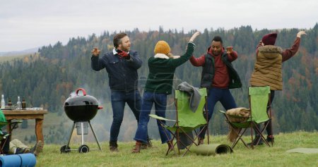 Téléchargez les photos : Groupe de voyageurs s'amusent pendant le barbecue sur la colline. Heureux amis multiethniques dansent se reposant à l'extérieur pendant le voyage de vacances. Montagnes paysage et forêt en arrière-plan. Amateurs de plein air. - en image libre de droit