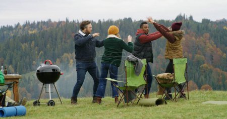 Photo for Group of travelers have fun during barbecue on hilltop. Happy multiethnic friends dance resting outdoors during vacation trip. Mountains landscape and forest in the background. Outdoor enthusiasts. - Royalty Free Image