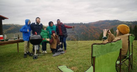 Photo for Multiethnic group of travelers stand near barbecue grill posing on camera. Woman in camp chair takes picture on phone. Big family resting outside of holiday house in the mountains. Tourism and travel. - Royalty Free Image