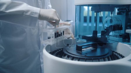 Téléchargez les photos : Homme scientifique met des échantillons de sang à partir de porte-tubes dans un dispositif automatique moderne pour les analyses biochimiques du sang. Un chimiste professionnel travaille dans un laboratoire de biotechnologie. Laboratoire de recherche avancée. - en image libre de droit