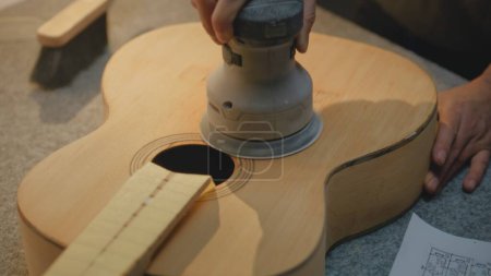 Téléchargez les photos : Gros plan du corps professionnel de la guitare de meulage de charpentier à l'aide de la rectifieuse. Artisan fabrique de la guitare acoustique. L'artisan travaille dans un atelier léger et élégant. Concept d'artisanat et de petite entreprise. - en image libre de droit