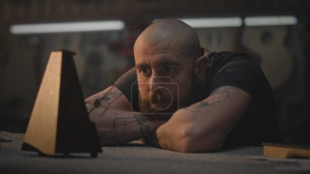 Photo for Middle aged tattooed craftsman sits at table in dark workroom. Professional Caucasian carpenter looks at metronome and rests after long working day in workshop. Handcraft and small business concept. - Royalty Free Image