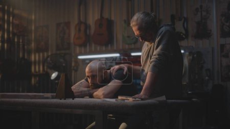 Foto de Trabajador cansado mira metrónomo, se sienta en taller moderno para la fabricación de instrumentos musicales. Maduro carpintero viene y pone la mano en colega de nuevo apoyándolo. Guitarras de madera hechas a mano cuelgan en la pared. - Imagen libre de derechos