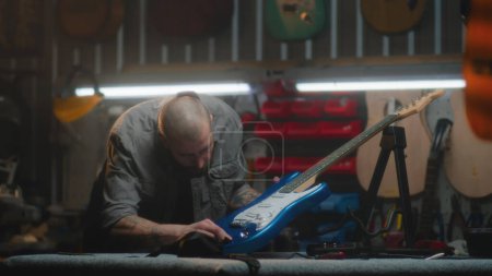 Photo for Male carpenter inspects guitar in detail and wipes with it rag. Craftsman makes wooden musical instrument in modern workshop. Colleague works in the background. Handicraft and small business concept. - Royalty Free Image