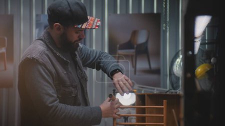 Téléchargez les photos : Menuisier mâle assemble table de chevet en bois sous lampe LED en utilisant un outil professionnel. Artisanat professionnel, l'artisan travaille dans un atelier moderne de fabrication de meubles. Artisanat et petite entreprise. - en image libre de droit