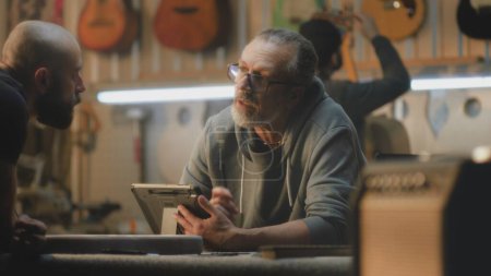 Téléchargez les photos : Homme artisan mature utilise tablette numérique pour dessiner des plans de guitare. Moyen âge tatoué charpentier parle avec un collègue, prend des manches de guitare. Les artisans travaillent dans un atelier élégant et léger. - en image libre de droit