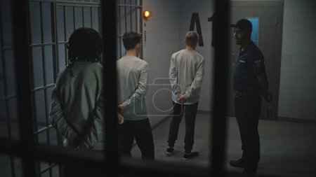 Foto de Un grupo de prisioneros está cerca de la celda de la cárcel, el alcaide habla con ellos. Adolescentes multiétnicos cumplen condena de prisión en un centro de detención. Jóvenes criminales en prisión. Sistema de justicia. Vista a través de barras de metal. - Imagen libre de derechos