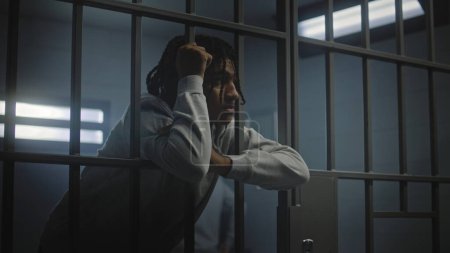 Upset African American teenager with face tattoos stands in prison cell in jail or youth detention center leaning on metal bars. Prison officer passes by young criminal or prisoner in the foreground.