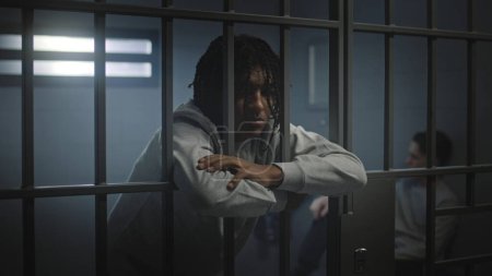 Depressed African American teenager with tattoos stands in prison cell in jail and looks at camera. Young inmate serves imprisonment term for crime. Juvenile detention center or correctional facility.