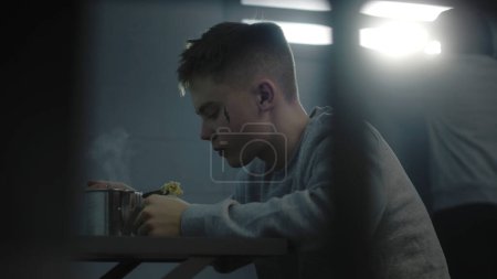 Téléchargez les photos : Un adolescent caucasien mange de la nourriture de prison à table dans sa cellule. Un jeune criminel purge une peine d'emprisonnement pour crime dans un centre de détention pour jeunes. Un autre détenu regarde la fenêtre en arrière-plan. - en image libre de droit