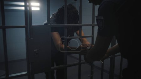 Warden brings new prisoner in jail cell and takes off his handcuffs. African American teen serves imprisonment term in correctional facility or detention center. Young inmates in jail. Tracking shot.