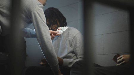 Foto de Tres prisioneros adolescentes multiétnicos juegan cartas en la celda de la prisión. Los reclusos jóvenes cumplen condena de prisión por delitos en la cárcel. Centro de detención juvenil o correccional. Vista a través de barras de metal. - Imagen libre de derechos