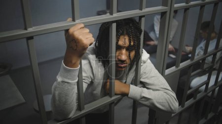 Wütender afroamerikanischer Teenager steht in einer Gefängniszelle im Gefängnis, hält Metallstangen in der Hand. Im Hintergrund spielen junge Häftlinge Karten auf dem Bett. Jugendhaftanstalt oder Justizvollzugsanstalt. Hoher Winkel.