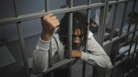 Angry African American teenage prisoner stands in prison cell in jail, holds metal bars. Young inmates play cards on bed in the background. Youth detention center or correctional facility. High angle.